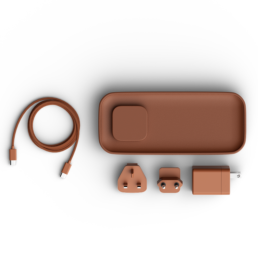 A set of travel charging accessories in a matte terracotta brown color, including a coiled USB-C to USB-C cable, a rectangular charging dock, a UK three-pin plug adapter, a USB-C to USB-C adapter, and a wall charger, all arranged neatly on a white background.