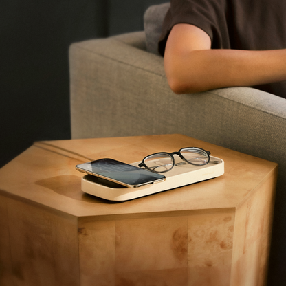 A beige rectangular wireless charging tray resting on a light wood hexagonal side table, holding a smartphone and eyeglasses, with a person seated in a gray armchair in the background.